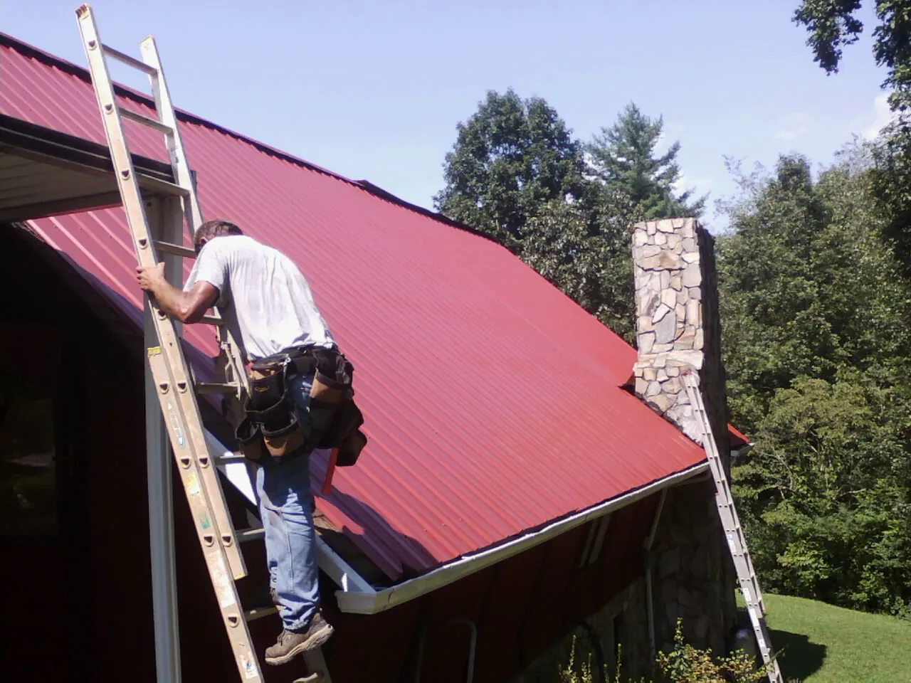 Roofer on a ladder beside a red metal roof and stone chimney — Soffit & Fascia Repair by Roofing Brothers in Novelty